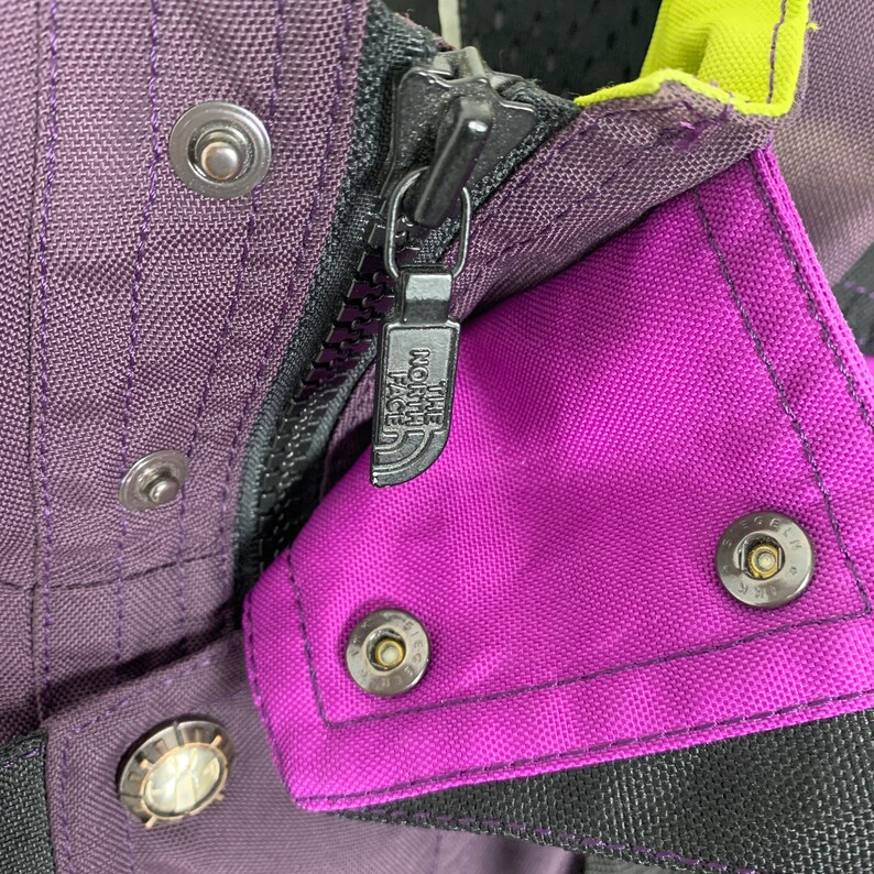 May include: Close-up of a purple and black jacket zipper with the The North Face logo. The zipper is metal and has a black pull tab. The jacket is made of a durable nylon fabric.