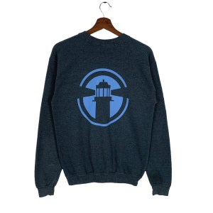 Peut inclure: Sweat-shirt gris foncé avec un motif de phare bleu clair dans un cercle. Le sweat-shirt à col rond et à manches longues est suspendu à un cintre en bois sur fond blanc.