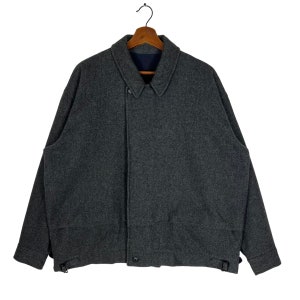 May include: A gray wool jacket with a button closure and a collar. The jacket has two pockets on the front and a strap on each side.