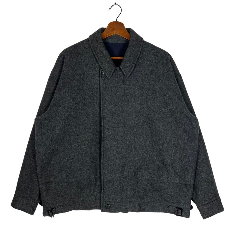 May include: A gray wool jacket with a button closure and a collar. The jacket has two pockets on the front and a strap on each side.