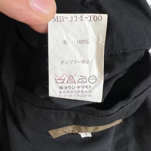Op de afbeelding: Close-up van een zwart kledingstuk met een wit label. Het label toont Japanse tekst, waaronder "毛 100%" en verzorgingssymbolen. Het label bevat ook de tekst "MB-J14-100".