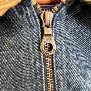 Pode incluir: Close-up de um z&iacute;per de jaqueta jeans azul com um puxador de cor lat&atilde;o. O puxador &eacute; gravado com o nome da marca "Pendleton" e um logotipo. A jaqueta tem uma gola de veludo cotel&ecirc;.