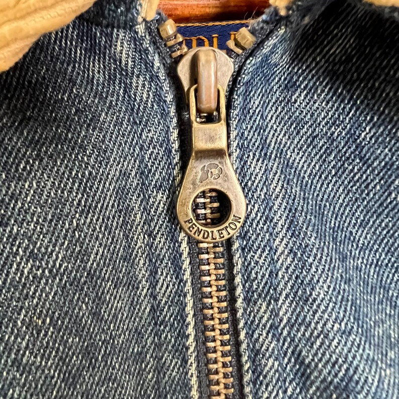 Pode incluir: Close-up de um z&iacute;per de jaqueta jeans azul com um puxador de cor lat&atilde;o. O puxador &eacute; gravado com o nome da marca "Pendleton" e um logotipo. A jaqueta tem uma gola de veludo cotel&ecirc;.