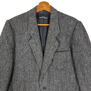 Puede incluir: Un blazer gris con dise&ntilde;o de espiga, solapas con muescas, cierre de un solo bot&oacute;n y bolsillo en el pecho. El blazer est&aacute; colgado de una percha de madera. La etiqueta dice "tricot COMME des GAR&Ccedil;ONS".