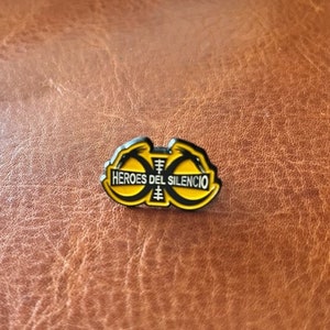 May include: A yellow and black enamel pin with the text "HEROES DEL SILENCIO" in white. The pin has a unique design with two stylized figures. The pin is set against a brown leather background.