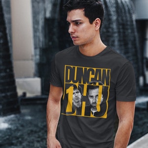 May include: A dark gray t-shirt with a yellow graphic print of the text "Duncan 1 DH3" and two black and white images of men's faces.