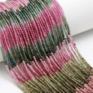 May include: A collection of small, multi-coloured beads in pink, green, and brown. The beads are strung together, creating a layered effect. The strands are draped over a white surface, with white tassels at the ends. The beads appear to be gemstones.