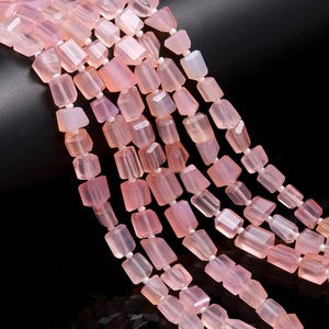 Puede incluir: Múltiples hebras de cuentas de piedras preciosas de color rosa pálido con forma rectangular. Las cuentas están ensartadas, formando varias hebras. Los extremos de las hebras están adornados con borlas. El fondo es negro.