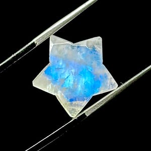 May include: A star-shaped gemstone with a blue and white iridescent glow, held by silver tweezers against a black background. The gemstone's facets catch the light, creating a shimmering effect.