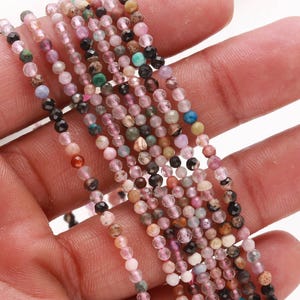 May include: Strands of small, multi-colored gemstone beads. The beads are a mix of pink, green, black, and brown, with a faceted surface. The beads are held in a hand, suggesting they are ready for jewelry making.