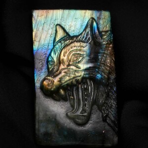 Könnte beinhalten: Ein rechteckiger Labradoritstein, der mit einem detaillierten Wolfskopf geschnitzt wurde. Der Wolf wird mit offenem Maul dargestellt, wobei die Zähne zu sehen sind. Der Stein hat schillernde Farben von Blau, Grün und Gold.