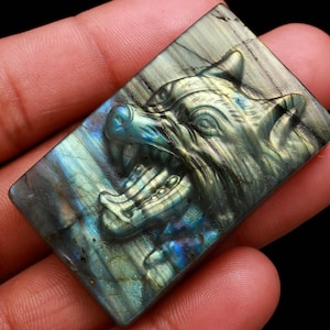 Könnte beinhalten: Ein rechteckiger Labradoritstein, der mit einem detaillierten Wolfskopf geschnitzt wurde. Die Schnitzerei zeigt das offene Maul des Wolfs, seine Zähne und Gesichtsdetails. Der Stein hat eine Mischung aus blauen, grünen und grauen Farben.