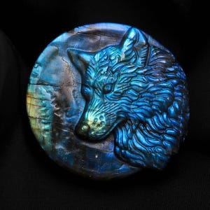 Könnte beinhalten: Ein runder, geschnitzter Stein mit einem detaillierten Wolfskopf in Blau- und Türkistönen. Der Stein hat eine polierte Oberfläche und ist vor einem schwarzen Hintergrund platziert. Das Fell des Wolfes ist aufwendig geschnitzt.