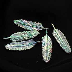 Peut inclure: Cinq pendentifs en forme de plumes iridescentes avec une surface chatoyante multicolore. Les plumes présentent un motif texturé et détaillé sur fond noir. Les pendentifs sont probablement destinés à la fabrication de bijoux ou à des projets d'artisanat.