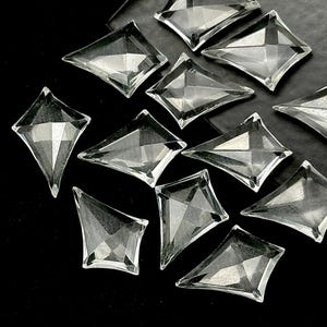 May include: Clear, faceted glass diamond-shaped embellishments scattered on a black surface. The embellishments have a reflective quality, catching the light and creating a sparkling effect. These decorative pieces are ideal for craft projects.