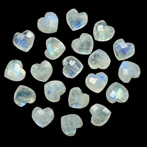 May include: A collection of faceted, heart-shaped moonstone gemstones. The stones are a translucent white with iridescent blue and yellow flashes, set against a black background. Ideal for jewelry making or crafts.