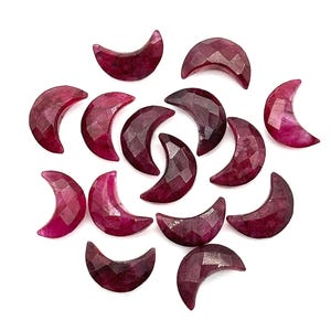 May include: A collection of ruby crescent moon gemstones, each with a faceted surface. The gems vary in shades of deep red and pink, scattered against a white background. These polished stones are ideal for jewelry making.