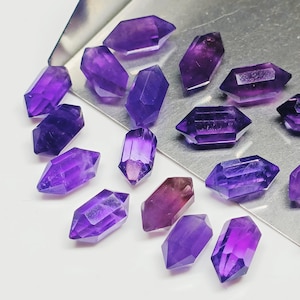 May include: A collection of 14 amethyst crystals, each cut into a double-pointed shape. The crystals are various shades of purple and are arranged on a silver tray.