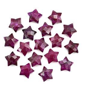 Puede incluir: Una colección de 17 piedras preciosas de rubí en forma de estrella facetadas. Las piedras de rubí son de color rojo intenso con un tono púrpura. Las piedras están dispuestas en círculo sobre un fondo blanco.