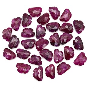 May include: A collection of faceted, deep red ruby gemstones. Each stone has a unique, cloud-like shape with multiple facets, creating a sparkling effect. The rubies are scattered on a white surface, showcasing their rich color and intricate cuts.