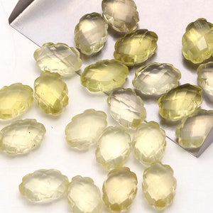 Pode incluir: Uma coleção de contas de pedras preciosas ovaladas facetadas de cor amarelo claro. As contas estão espalhadas sobre uma superfície branca e uma pá de metal.