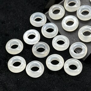 May include: A collection of translucent, donut-shaped beads. The beads are a pale, milky white color and have a smooth, polished appearance. They are arranged on a dark, textured surface.