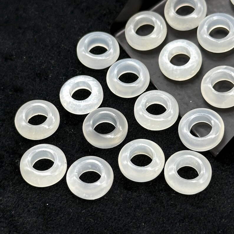 May include: A collection of translucent, donut-shaped beads. The beads are a pale, milky white color and have a smooth, polished appearance. They are arranged on a dark, textured surface.