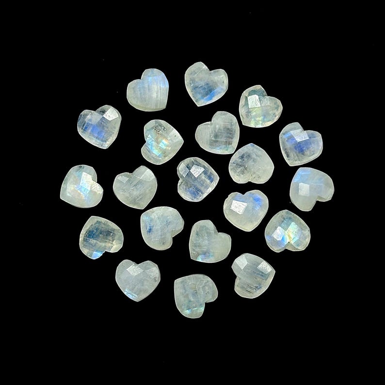 May include: A collection of faceted, heart-shaped moonstone gemstones. The stones are a translucent white with iridescent blue and yellow flashes, set against a black background. Ideal for jewelry making or crafts.
