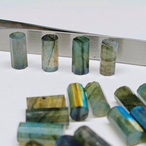 May include: A collection of iridescent green and blue labradorite gemstone beads in various shapes and sizes. Some beads are cylindrical, while others are rectangular. The beads are arranged on a white surface.