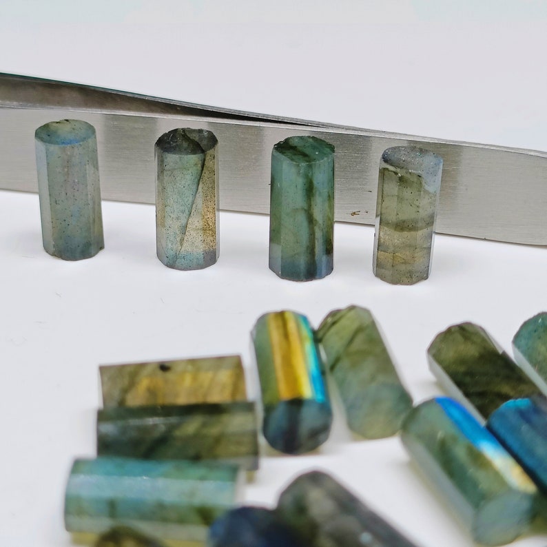 May include: A collection of iridescent green and blue labradorite gemstone beads in various shapes and sizes. Some beads are cylindrical, while others are rectangular. The beads are arranged on a white surface.
