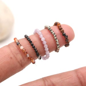 May include: Four beaded rings in different colors and materials. The rings are stacked on a finger, showing the different styles. The rings are made of amethyst, black onyx, tiger's eye, and pyrite.