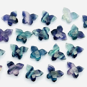 Può includere: Una collezione di 18 perline di pietra preziosa a forma di farfalla, ciascuna con una miscela unica di tonalità blu, verdi e viola. Le perline sono sfaccettate e hanno una finitura lucida.