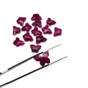 May include: Faceted, deep magenta butterfly-shaped gemstones. One gem is held by tweezers, showing its intricate cut. The gems are scattered on a white background, highlighting their colour and form.