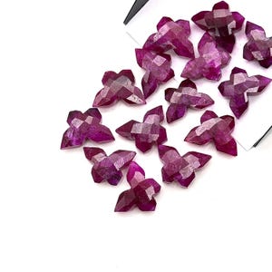 May include: A collection of faceted, deep magenta gemstone flower-shaped beads. The beads have a multi-faceted surface, catching the light and creating a sparkling effect. The beads are scattered on a white surface, with a few partially overlapping.