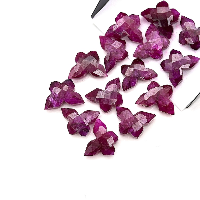 May include: A collection of faceted, deep magenta gemstone flower-shaped beads. The beads have a multi-faceted surface, catching the light and creating a sparkling effect. The beads are scattered on a white surface, with a few partially overlapping.