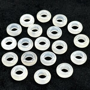 May include: A collection of translucent, off-white donut-shaped beads. Each bead has a circular hole in the center. The beads are scattered across a dark surface, creating a pattern of light and shadow.