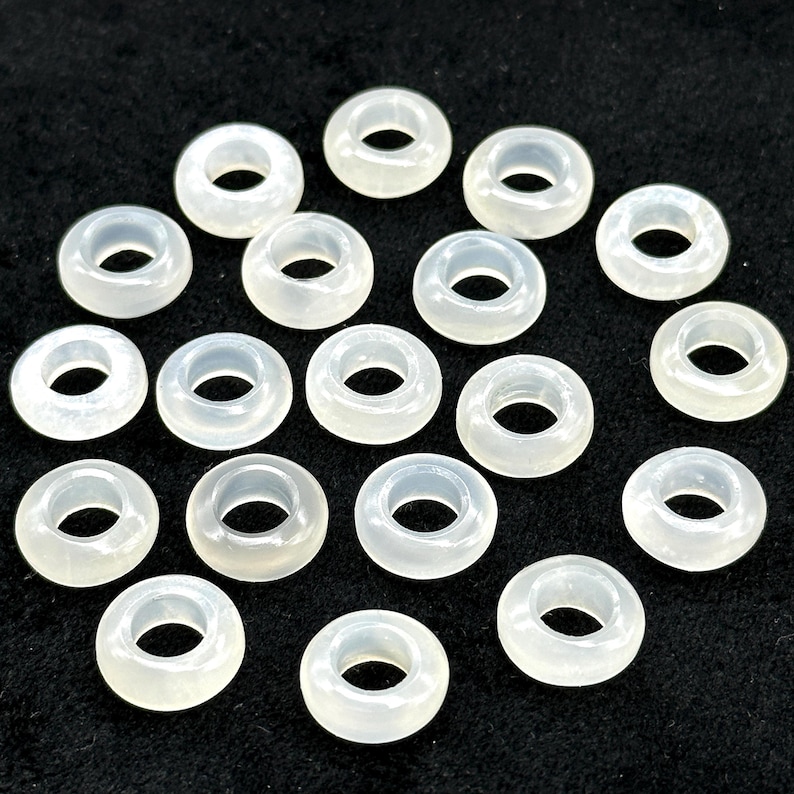 May include: A collection of translucent, off-white donut-shaped beads. Each bead has a circular hole in the center. The beads are scattered across a dark surface, creating a pattern of light and shadow.