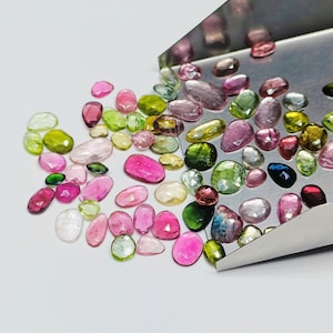Può includere: Una collezione di pietre preziose sciolte in varie tonalità di rosa, verde e blu. Le pietre preziose sono sparse su una superficie bianca e un vassoio di metallo.