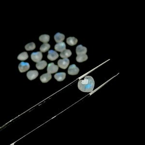 May include: A collection of heart-shaped moonstone gemstones. One gemstone is held by tweezers, showcasing its iridescent blue and white color. The other gemstones are scattered on a black background, highlighting their soft, translucent appearance.