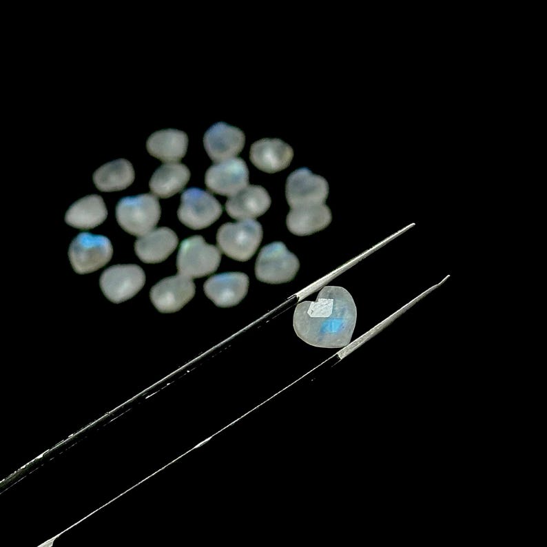 May include: A collection of heart-shaped moonstone gemstones. One gemstone is held by tweezers, showcasing its iridescent blue and white color. The other gemstones are scattered on a black background, highlighting their soft, translucent appearance.