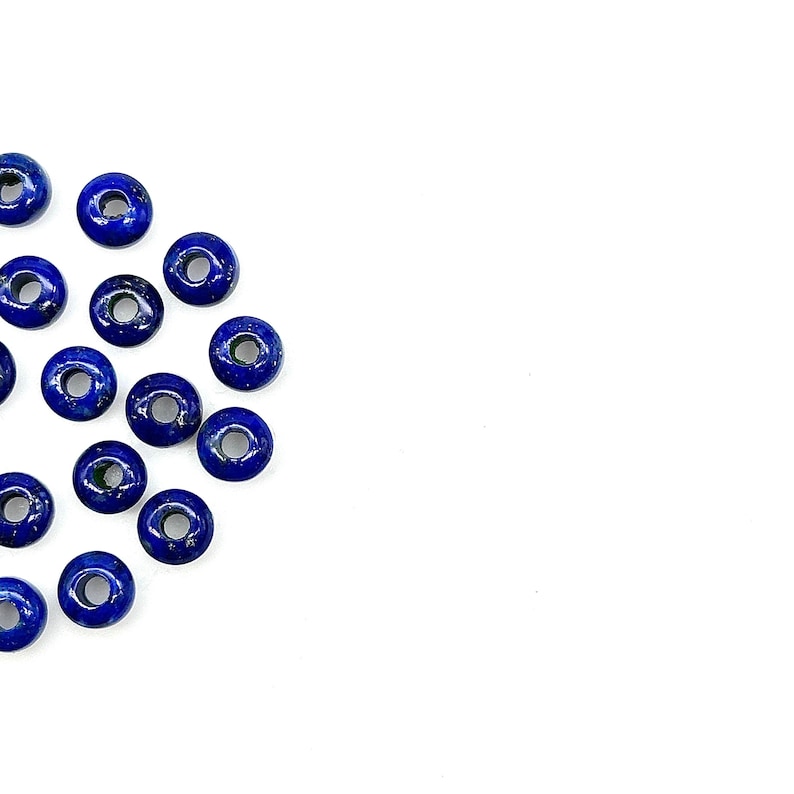 May include: A collection of round, blue lapis lazuli beads with a hole in the center. The beads are scattered on a white surface.