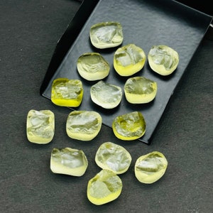 May include: A collection of 14 yellow-green, faceted, rough-cut gemstones. The stones are various sizes and shapes, and are arranged on a black surface.