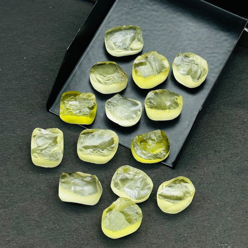 May include: A collection of 14 yellow-green, faceted, rough-cut gemstones. The stones are various sizes and shapes, and are arranged on a black surface.