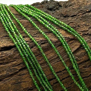 May include: A strand of green gemstone beads, likely peridot, arranged in parallel lines on a rough wooden surface. The beads are small and have a faceted, square shape.