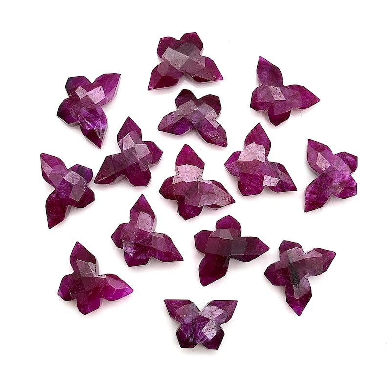 May include: A selection of faceted, butterfly-shaped gemstones in shades of deep magenta and purple. The gemstones are displayed on a white background, highlighting their intricate cuts and natural colour variations. These gemstones are suitable for jewellery making.
