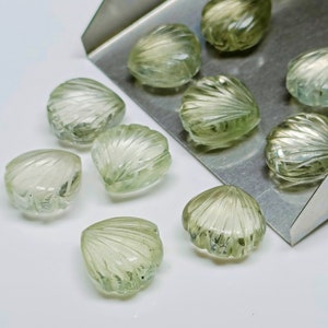 May include: A collection of ten green gemstone cabochons carved in the shape of seashells. The cabochons are arranged on a silver tray and a white surface.