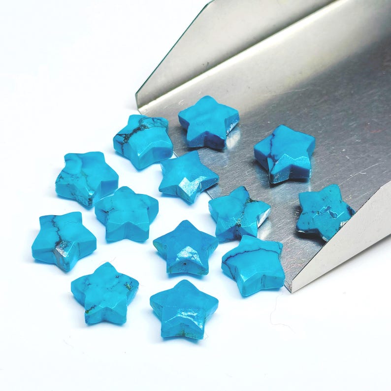 May include: A selection of turquoise star-shaped gemstones. The stones are faceted and a vibrant blue with dark veining. They are arranged on a white surface with a silver tool in the background.