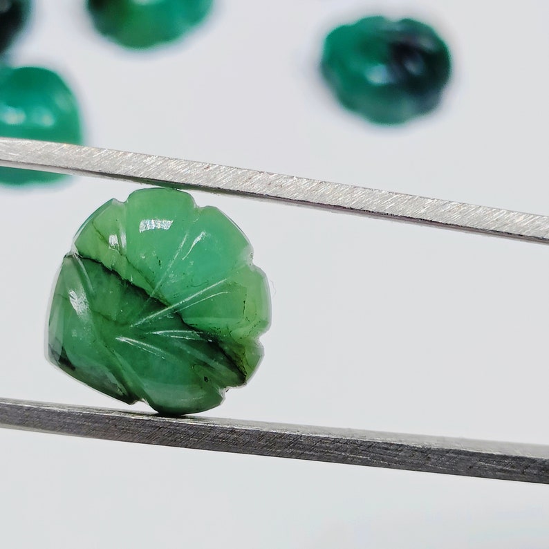 Puede incluir: Una piedra preciosa de esmeralda verde tallada en forma de hoja, sostenida por unas pinzas sobre un fondo blanco.