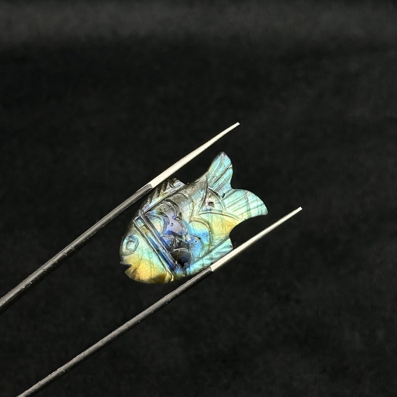 Puede incluir: Un colgante de pez de labradorita tallado con colores iridiscentes azules, verdes y dorados. El colgante est&aacute; sujeto por unas pinzas sobre un fondo negro.