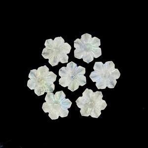 May include: Seven carved flower-shaped gemstones with a translucent, iridescent quality. The petals are detailed, and the gems are arranged against a black background. These could be used for jewelry or other crafts.